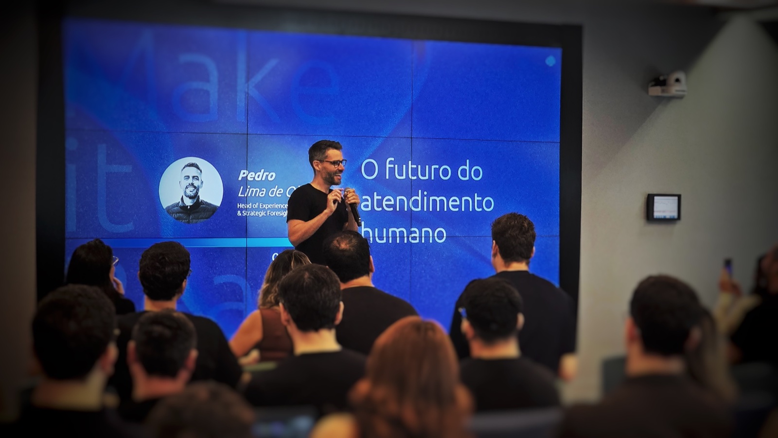 Pedro Lima apresentando no palco, com a plateia sentada à frente e uma tela atrás dele com o título &ldquo;O futuro do atendimento humano&rdquo;.